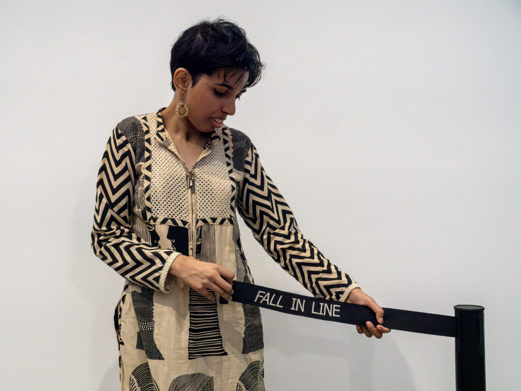 Priyanka d'Souza, vestindo um vestido preto e branco estampado, fica segurando um elemento de sua instalação de arte: uma torrente de controle de multidão cuja barreira lê "cair na fila" em todas as letras maiúsculas.