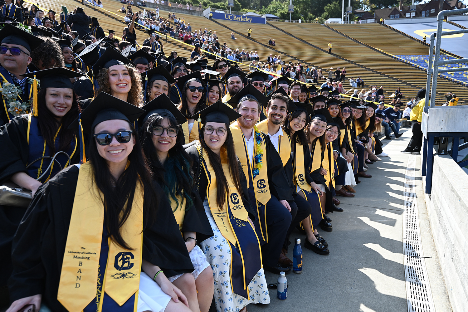 A long row of graduates seated in California Memorial Stadium smiles for the camera before the start of the 2025 campuswide commencement.