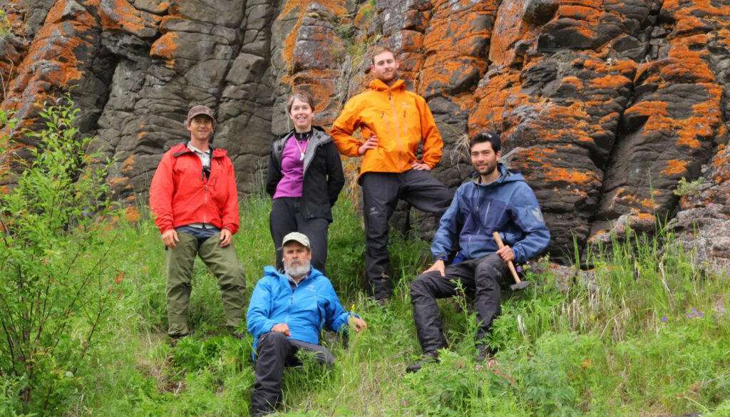 four men and one woman posing in front of an orange-streaked rock outcrop