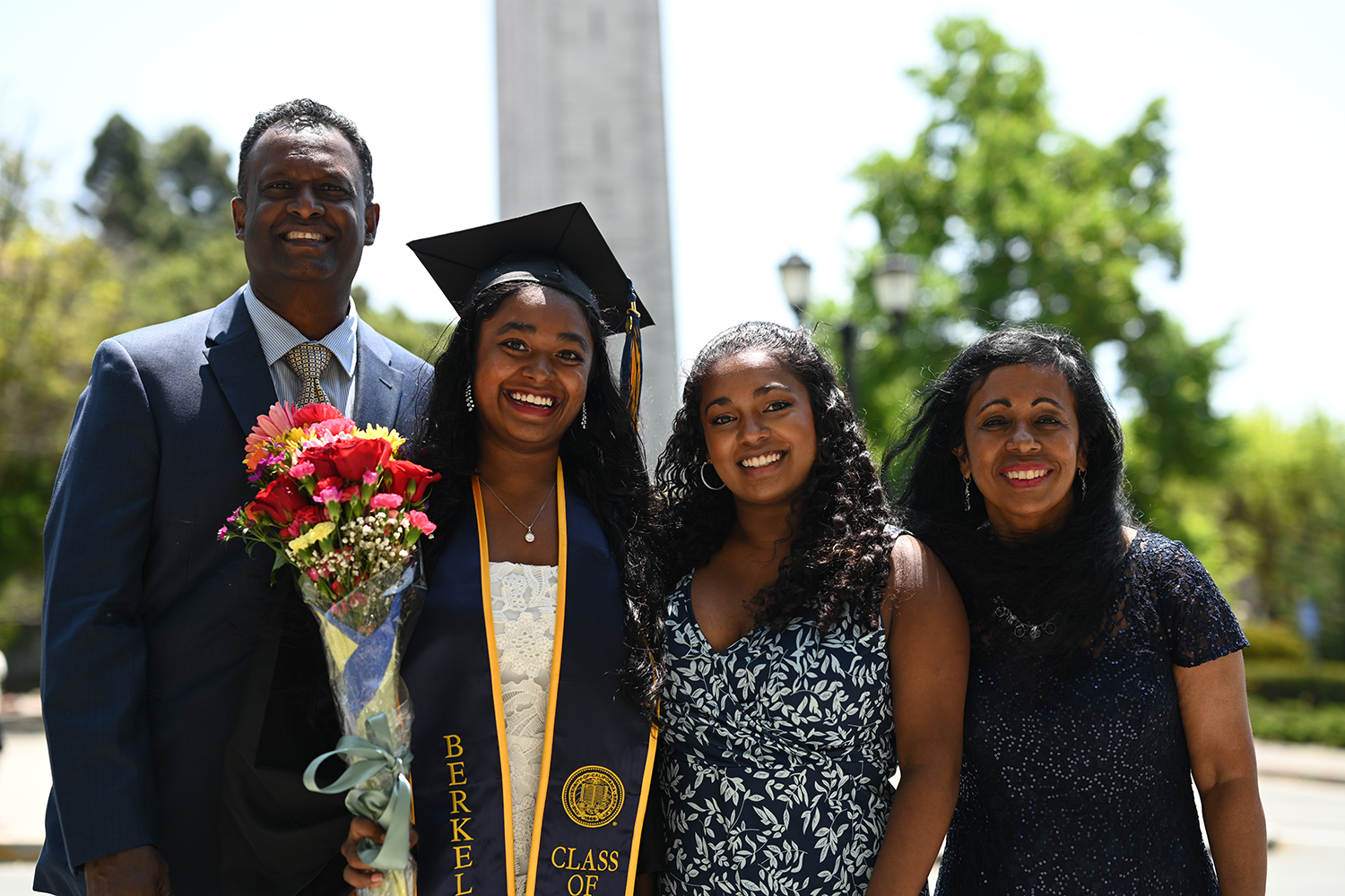 A family with a 2025 graduate poses with the Campanile in the distance. The grad is a young woman wearing regalia and holding a bouquet.