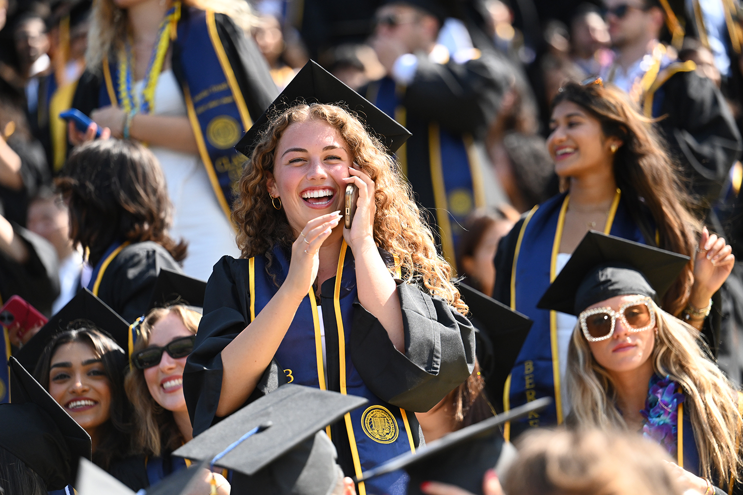 A graduate stands up in the bleachers at the 2025 campuswide commencement and smiles while talking on her phone.