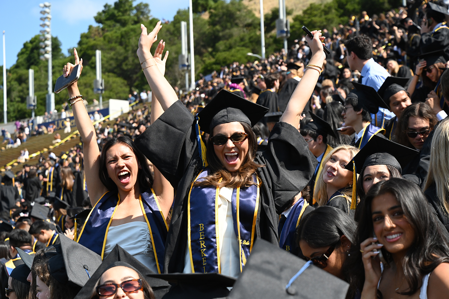 Two graduates at the 2025 campuswide commencement stand up in the crowd and throw up their arms into the air with joy.
