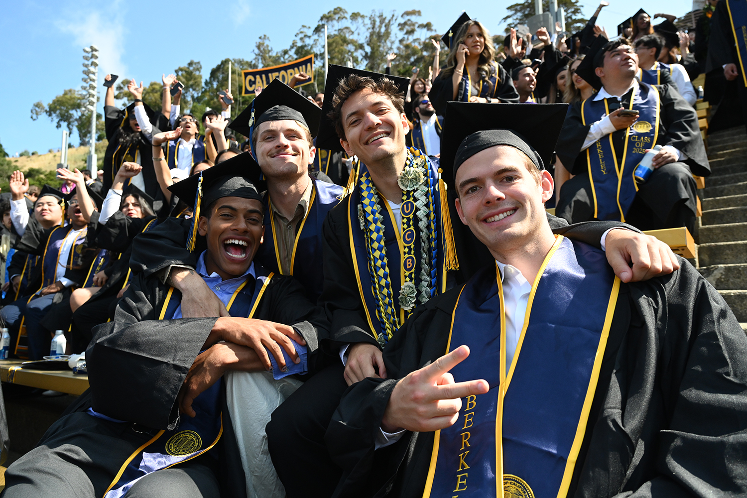 A group of four male students wearing regalia is all smiles and laughs as they sit in California Memorial Stadium waiting for commencement to start.