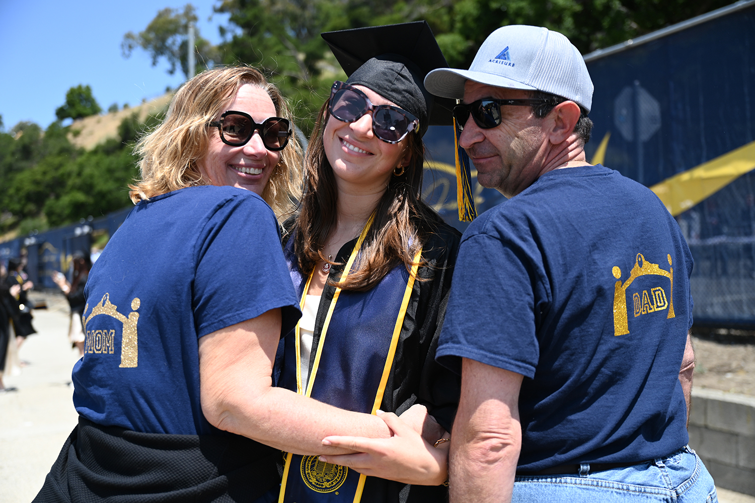 A mom, dad and graduating daughter all sport blue and gold at the 2025 campuswide commencement. The parents are wearing blue T-shirts with Mom and Dad on the back, plus the image of Sather Gate. The daughter is in regalia.
