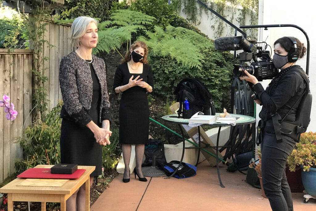 Nobel Prize winner Jennifer Doudna is in her backyard receiving her award during the COVID-19 pandemic because she can't get it in Sweden. She is in a black dress and tweed jacket with a string of pearls on. The Nobel medal is in a box on a table next to her, and two videographers from the UC Berkeley news office are filming the event.