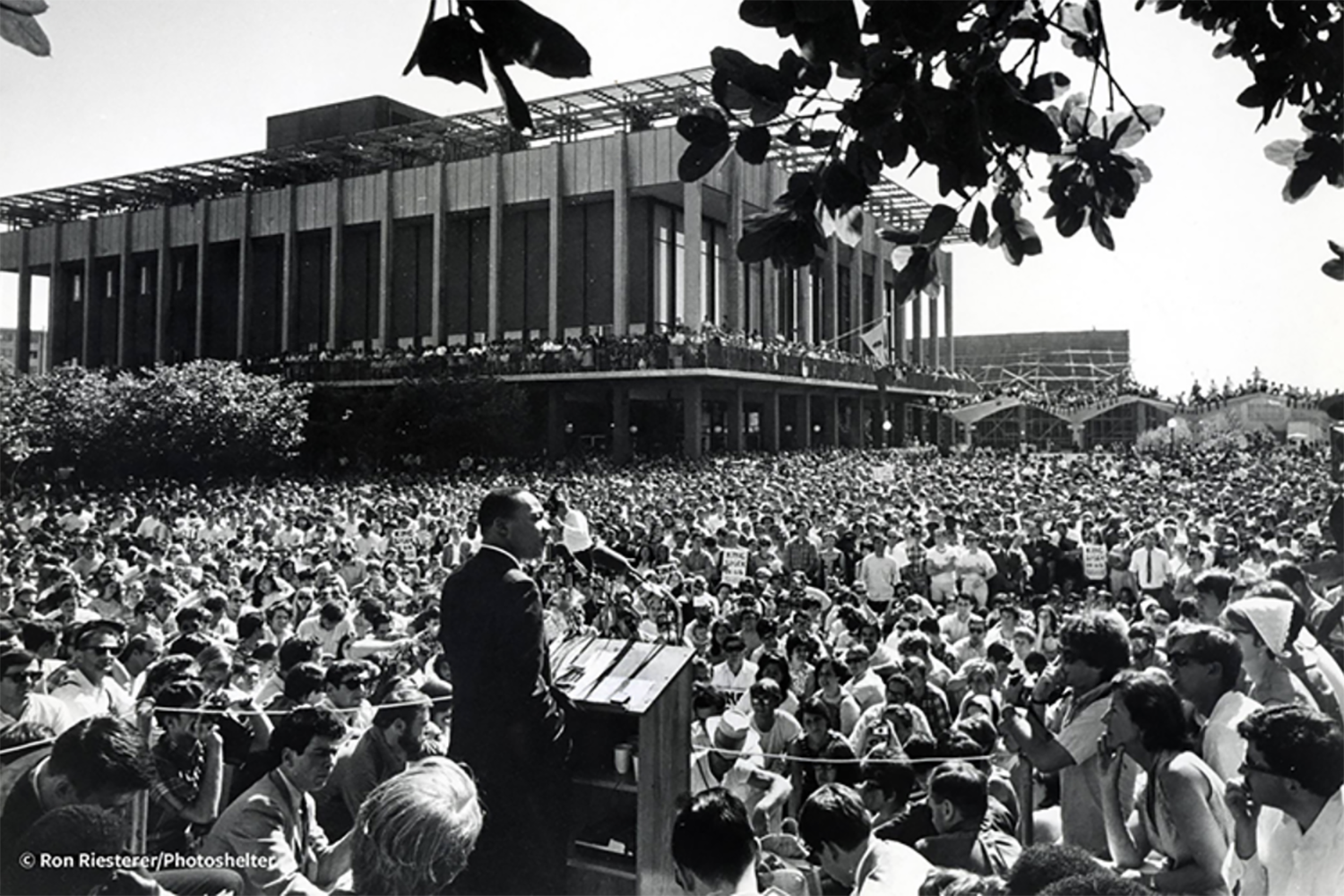 In this black and white photo, Dr. Martin Luther King Jr. speaks on Sproul Plaza before 7,000 people. He is facing a sea of students from behind a podium.