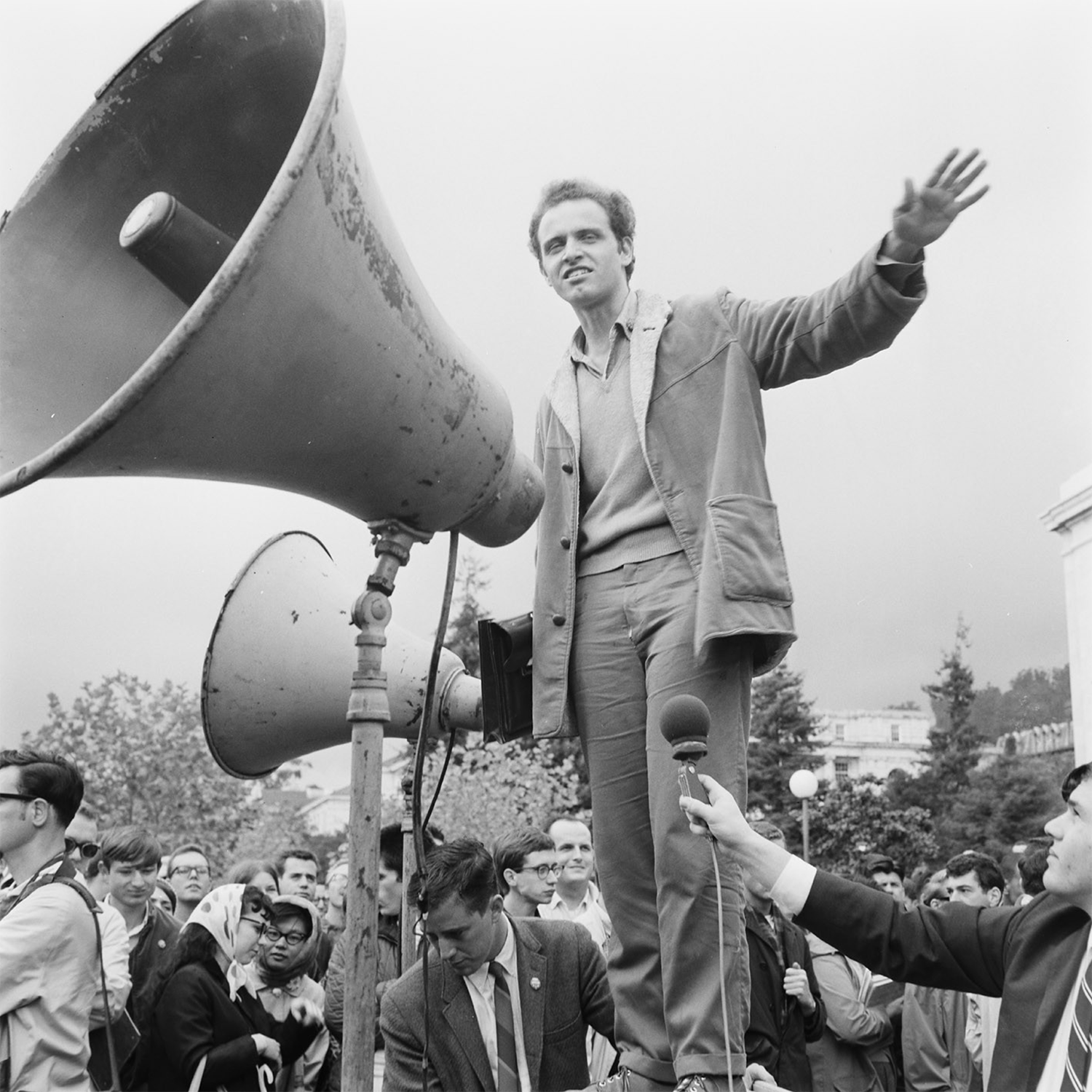Mario Savio, famous student activists, stands in the early 1960s on Sproul Hall next to giant speakers that project his words to the crowd, which includes a reporter with a microphone.