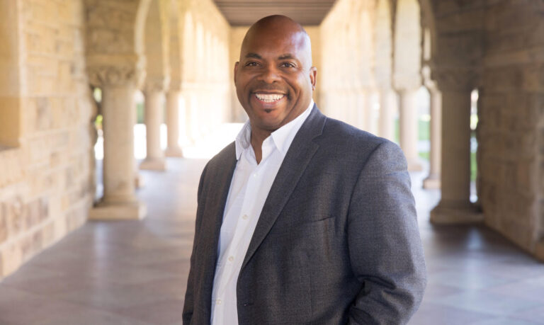 A bald Black man in a suit jacket and button-up smiles in an outdoor headshot