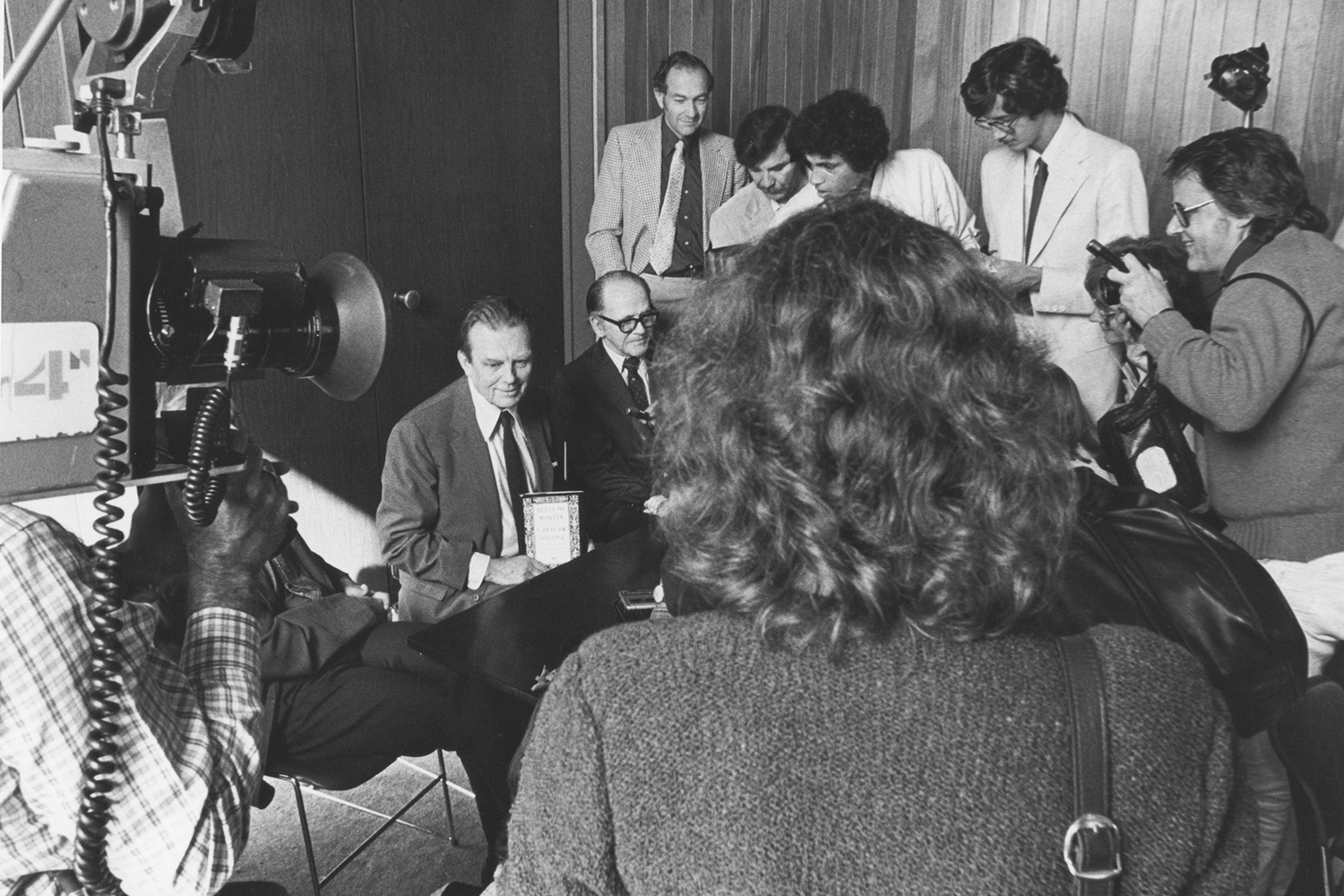 In this black and white photo, writer, poet and UC Berkeley professor Czeslaw Milosz speaks to reporters who surround him with cameras and microphones at a news conference arranged by the campus in honor of him winning the 1980 Nobel Prize in Literature. He is seated in a suit and tie holding a rectangular card in his hand.