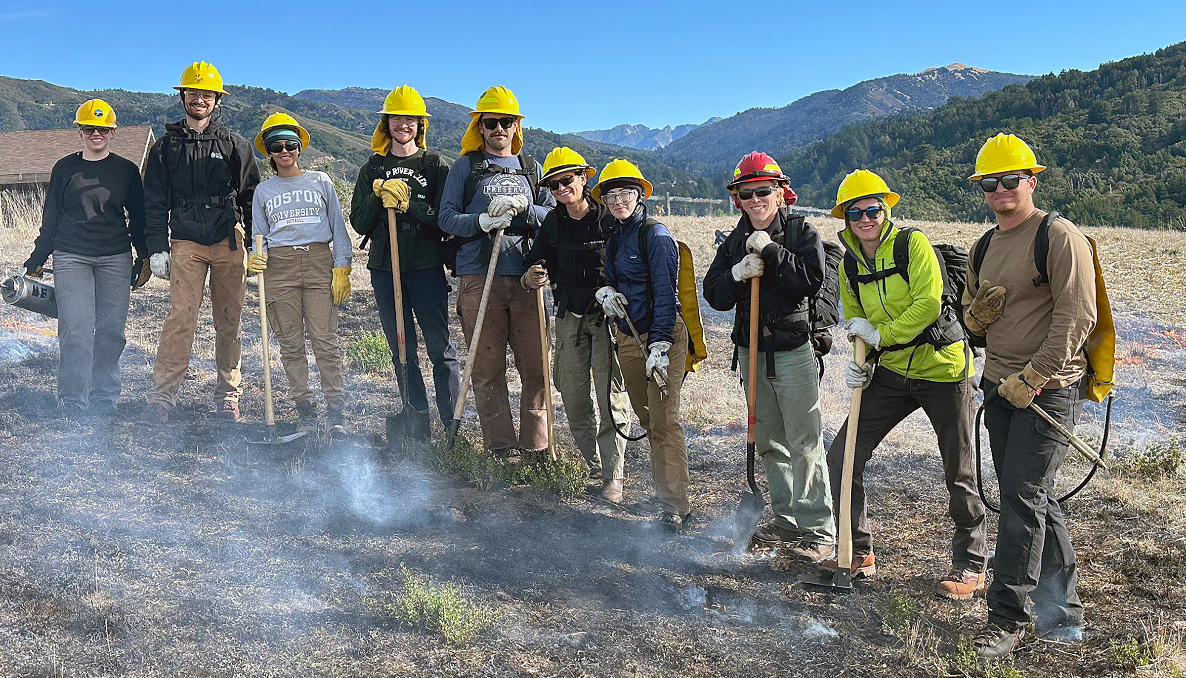 Workers in yellow hardhats, many holding shovels and other tools, stand in a smoky hillside field during a fire-suppression training.