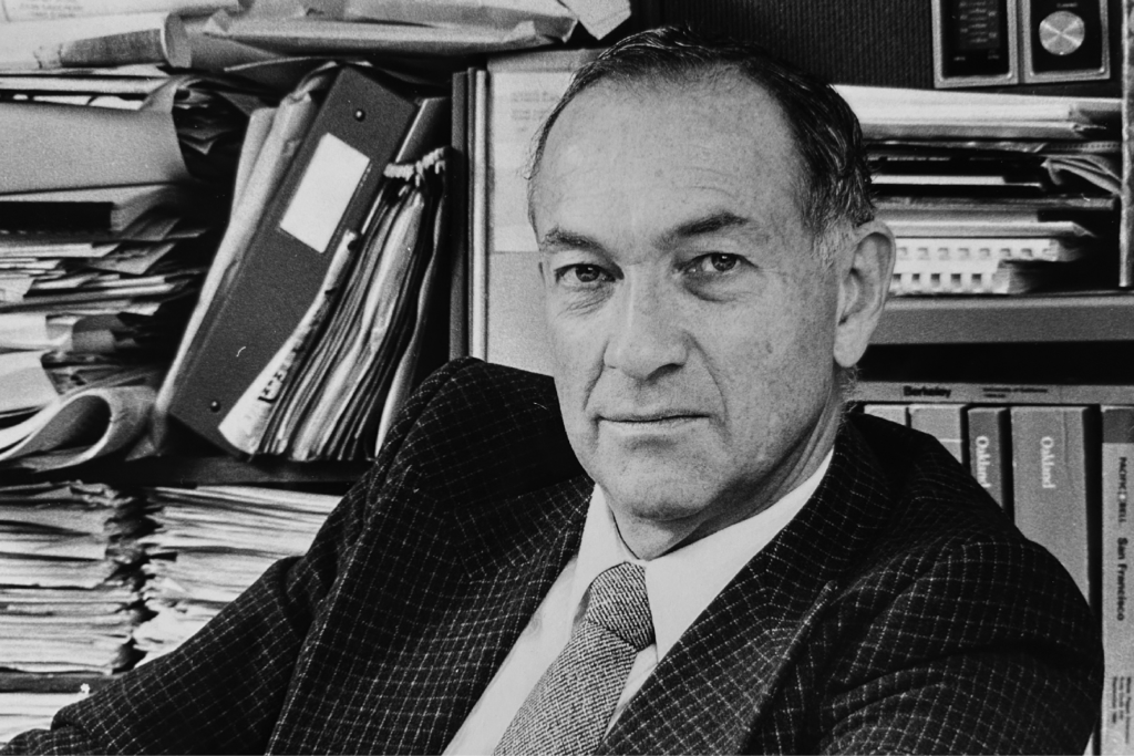 A black and white photo of Ray Colvig, who ran UC Berkeley's news bureau from 1972-1991. He's in a suit and tie and sitting in front of shelving that's crammed with papers and binders.