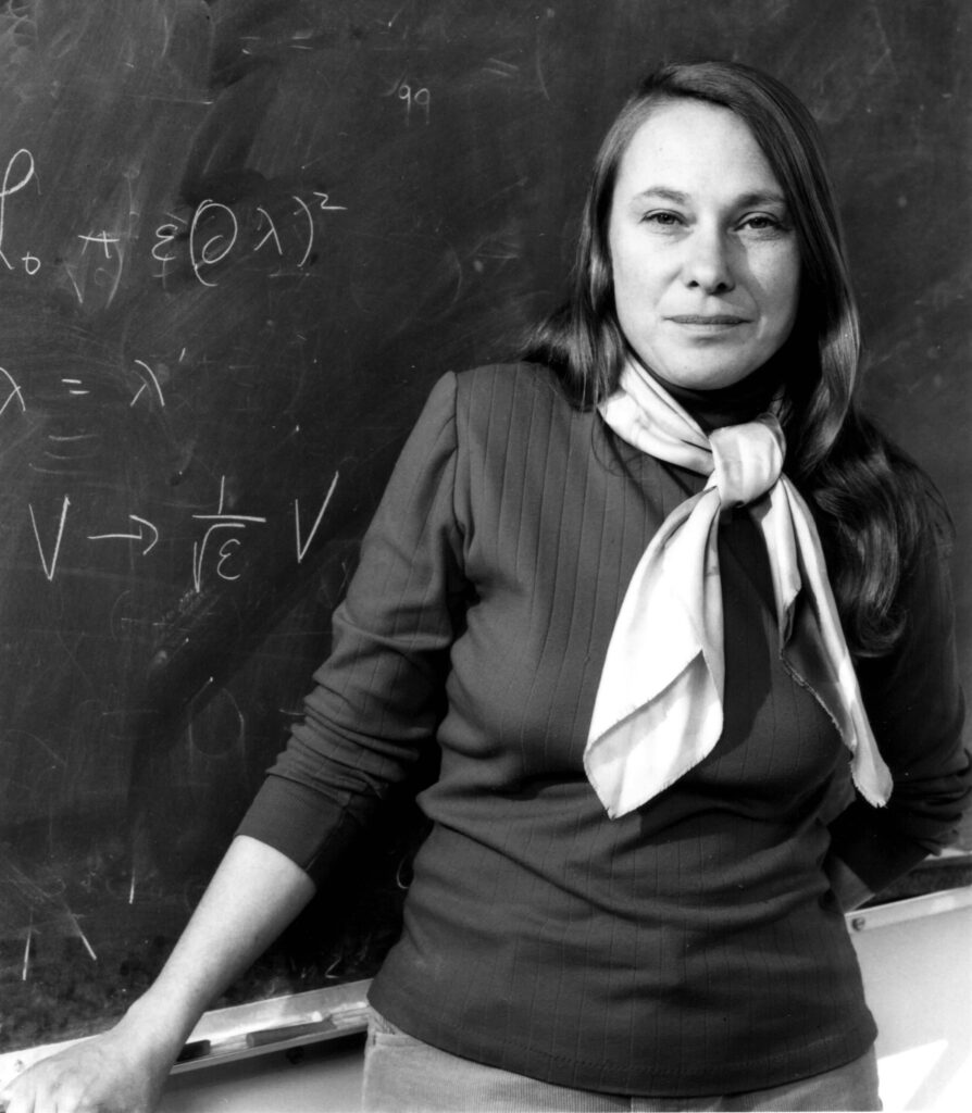 a woman wearing a dark blouse and light-colored scarf around her neck standing in front of a chalkboard