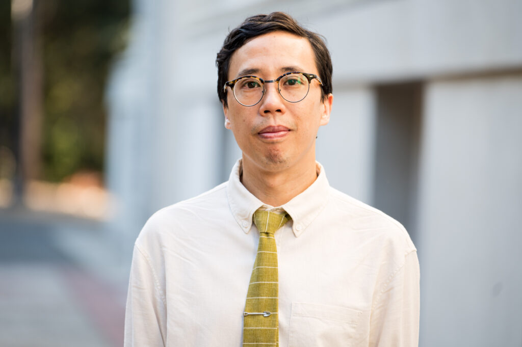 A headshot of an Asian American man, wearing glasses, a button-down and a tie, outside on Berkeley's campus