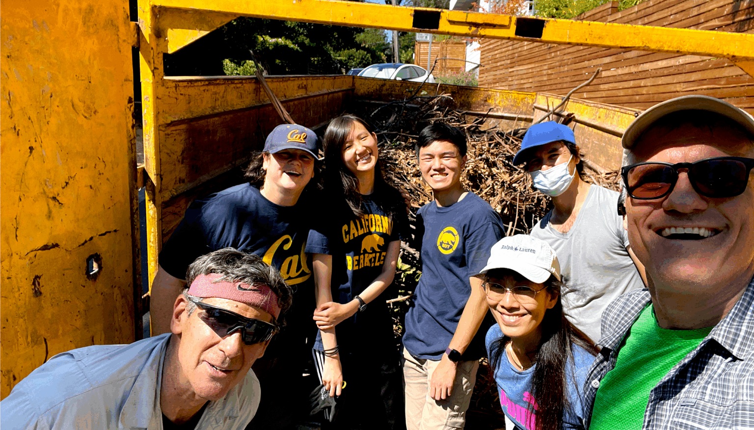 A group of seven people stands and smiles in front of a large yellow disposal bin that is filled with small twigs and branches. The bin is on a hilly street next to a white house.