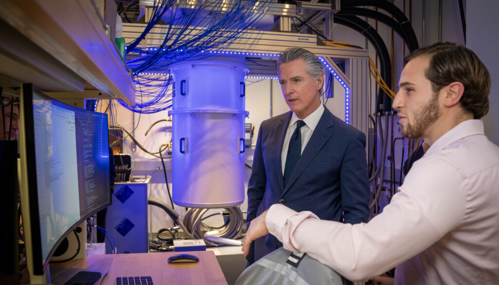 man in blue suit talking with a bearded guy in a white shirt, with a cylinder illuminated by blue light in the background
