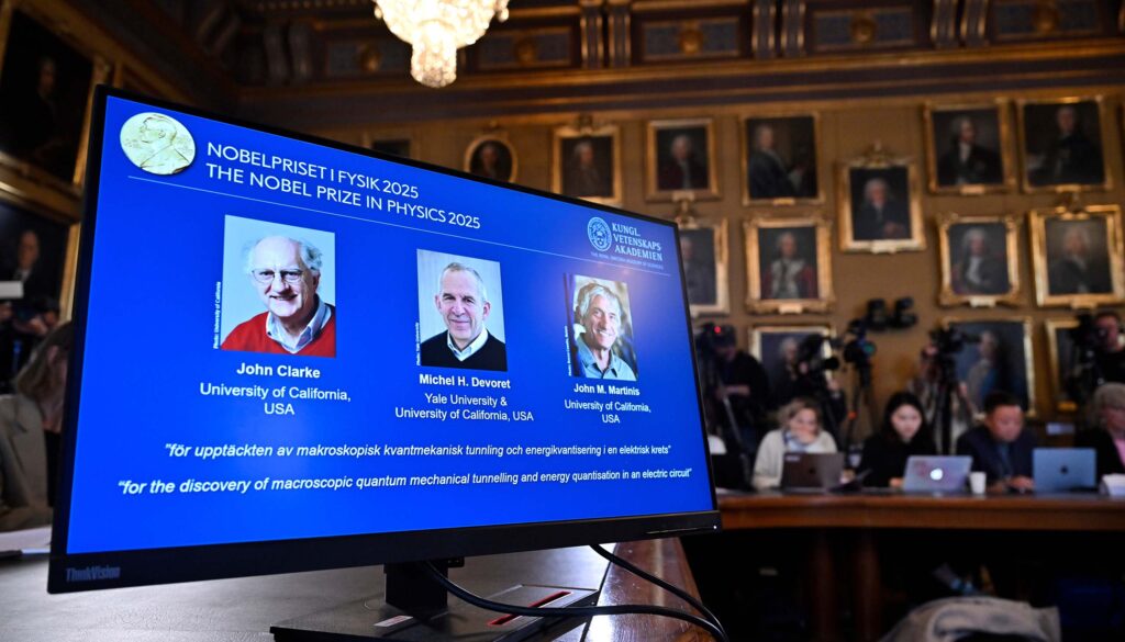 A screen shows the three 2025 Nobel laureates in Physics at the, with people sitting in the background.